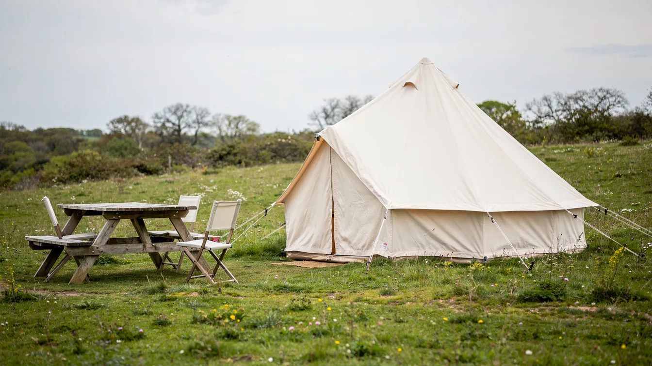 A simple canvas bell tent in a meadow with a modest picnic table and folding chairs